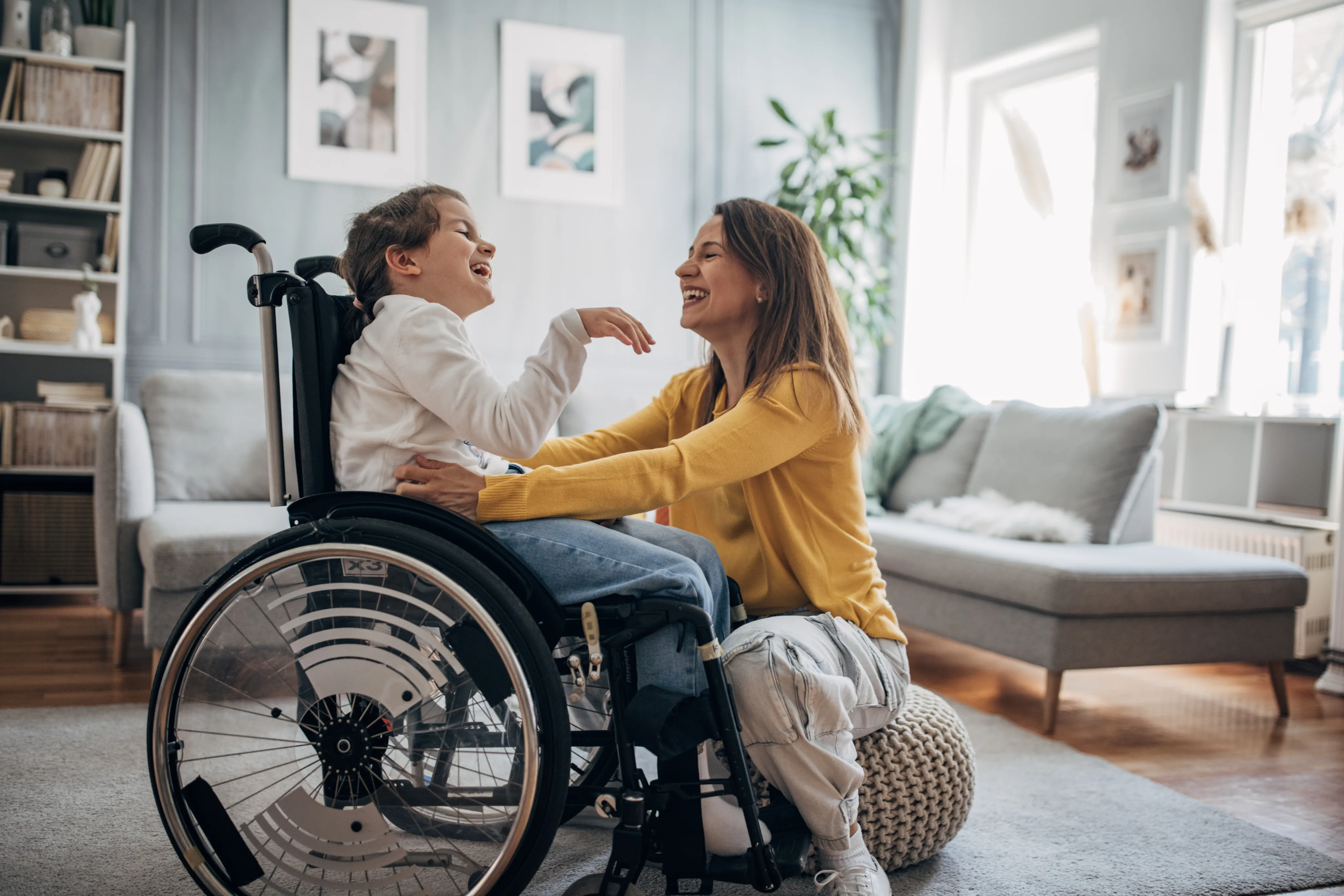 A caregiver laughs with a smiling child seated in a wheelchair at home.