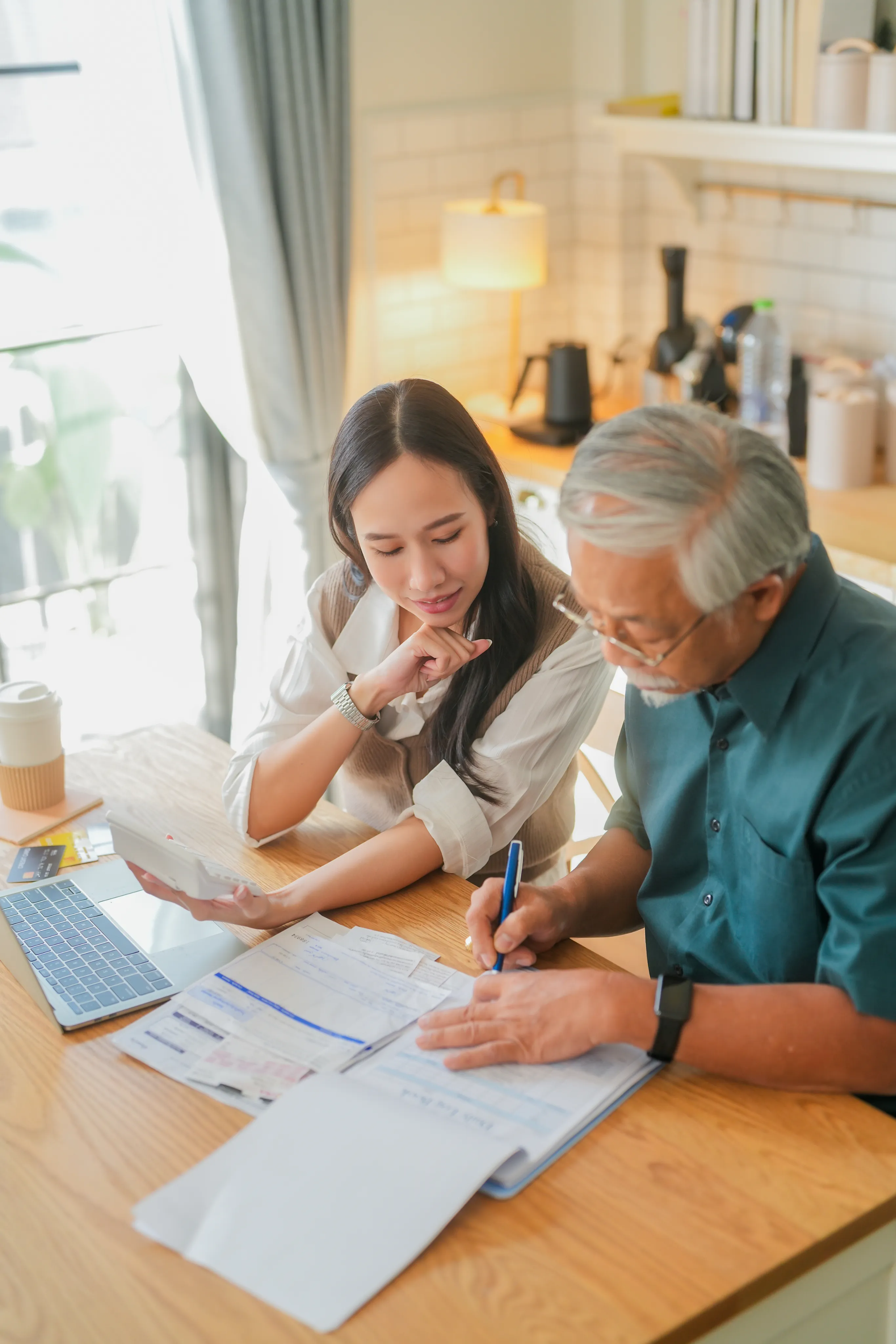 A woman helping an older man review and sign paperwork at a kitchen table
