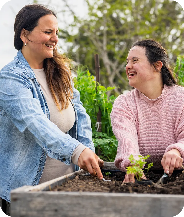 A caregiver and a young woman smiling together while planting seedlings in a raised garden bed