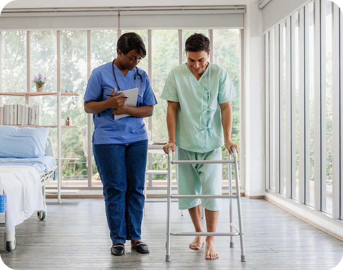 A nurse takes notes as a patient walks with the help of a walker in a hospital room