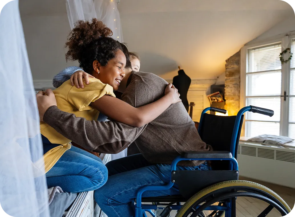 A smiling young woman hugs a man seated in a wheelchair at home